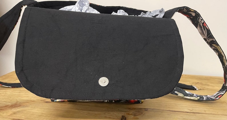 Puede incluir: Un bolso bandolera de tela negra con cierre de solapa y un bot&oacute;n a presi&oacute;n plateado. El bolso tiene un estampado floral en la parte posterior y una correa negra.