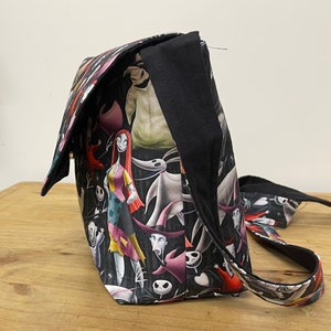 Puede incluir: Un bolso de hombro de tela negro y blanco con un estampado de Pesadilla Antes de Navidad con personajes como Jack Skellington, Sally y Oogie Boogie. El bolso tiene una correa negra.