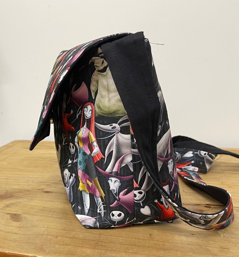 Puede incluir: Un bolso de hombro de tela negro y blanco con un estampado de Pesadilla Antes de Navidad con personajes como Jack Skellington, Sally y Oogie Boogie. El bolso tiene una correa negra.