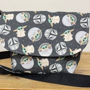May include: A black messenger bag with a repeating pattern of a green character and a Mandalorian helmet. The bag has a black adjustable strap.