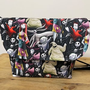 Puede incluir: Una bolsa de mensajero de lona negra con un estampado colorido de personajes de la pel&iacute;cula Pesadilla antes de Navidad. El estampado presenta a Jack Skellington, Sally, Oogie Boogie y otros personajes.