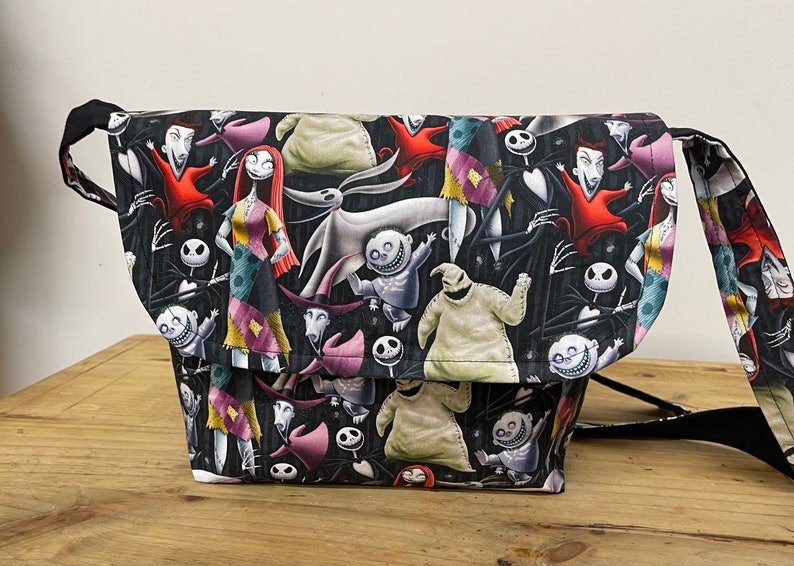 Puede incluir: Una bolsa de mensajero de lona negra con un estampado colorido de personajes de la pel&iacute;cula Pesadilla antes de Navidad. El estampado presenta a Jack Skellington, Sally, Oogie Boogie y otros personajes.