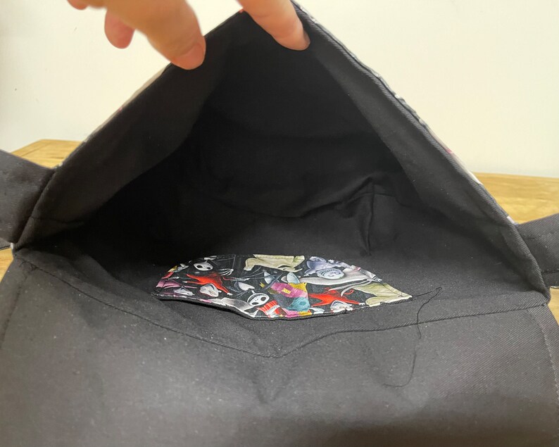 Puede incluir: Bolso tote de tela negro con un forro estampado en blanco y negro. El forro presenta una variedad de personajes y dise&ntilde;os.