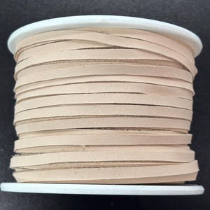 May include: A spool of flat, light beige leather cord wrapped neatly around a white plastic reel. The cord is thin and uniform in width, showing a smooth, matte texture across the multiple layers wound tightly around the central cylinder.