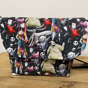 Puede incluir: Una bolsa de mensajero de lona negra con un estampado colorido de personajes de la pel&iacute;cula Pesadilla antes de Navidad. El estampado presenta a Jack Skellington, Sally, Oogie Boogie y otros personajes.