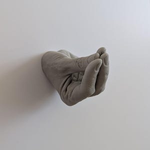May include: A gray concrete hand sculpture with fingers curled inward, resembling a gesture of offering or receiving.