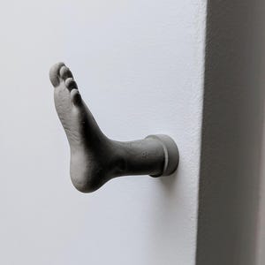 May include: A gray, foot-shaped wall hook made of concrete. The hook is mounted on a white wall and has a detailed texture, showing the toes and sole of the foot. The hook is designed to hold items.