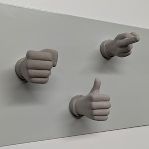以下が含まれることがあります： 壁に取り付けられた3Dプリントされた灰色の手3つ。1つの手は親指を立てており、もう1つは拳を握り、もう1つは右を指している。