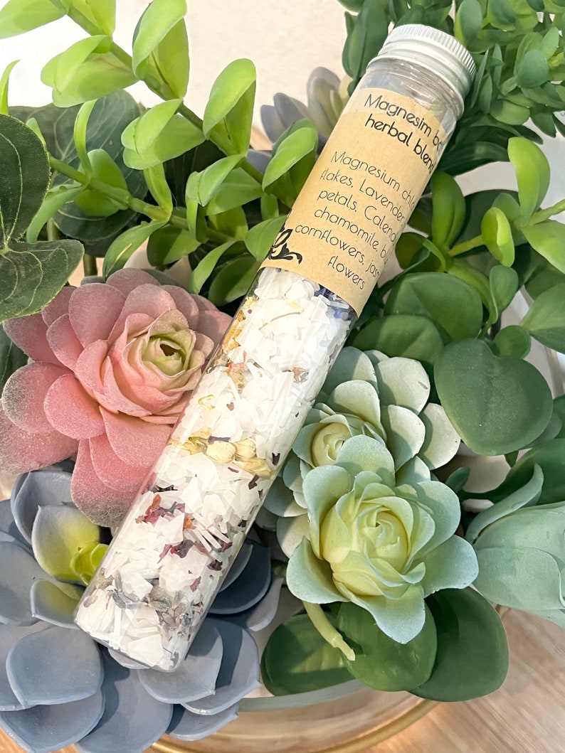 Puede incluir: Un tubo de vidrio lleno de una mezcla de sales de ba&ntilde;o blancas. La etiqueta dice "Magnesium bath herbal blend" y enumera los ingredientes: copos de cloruro de magnesio, p&eacute;talos de lavanda, p&eacute;talos de cal&eacute;ndula, manzanilla, aciano, flores de jazm&iacute;n.