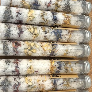 Puede incluir: Primer plano de varios tubos de vidrio transparente llenos de sales de ba&ntilde;o blancas y p&eacute;talos de flores secas. Los tubos tienen tapas plateadas y est&aacute;n dispuestos en fila.