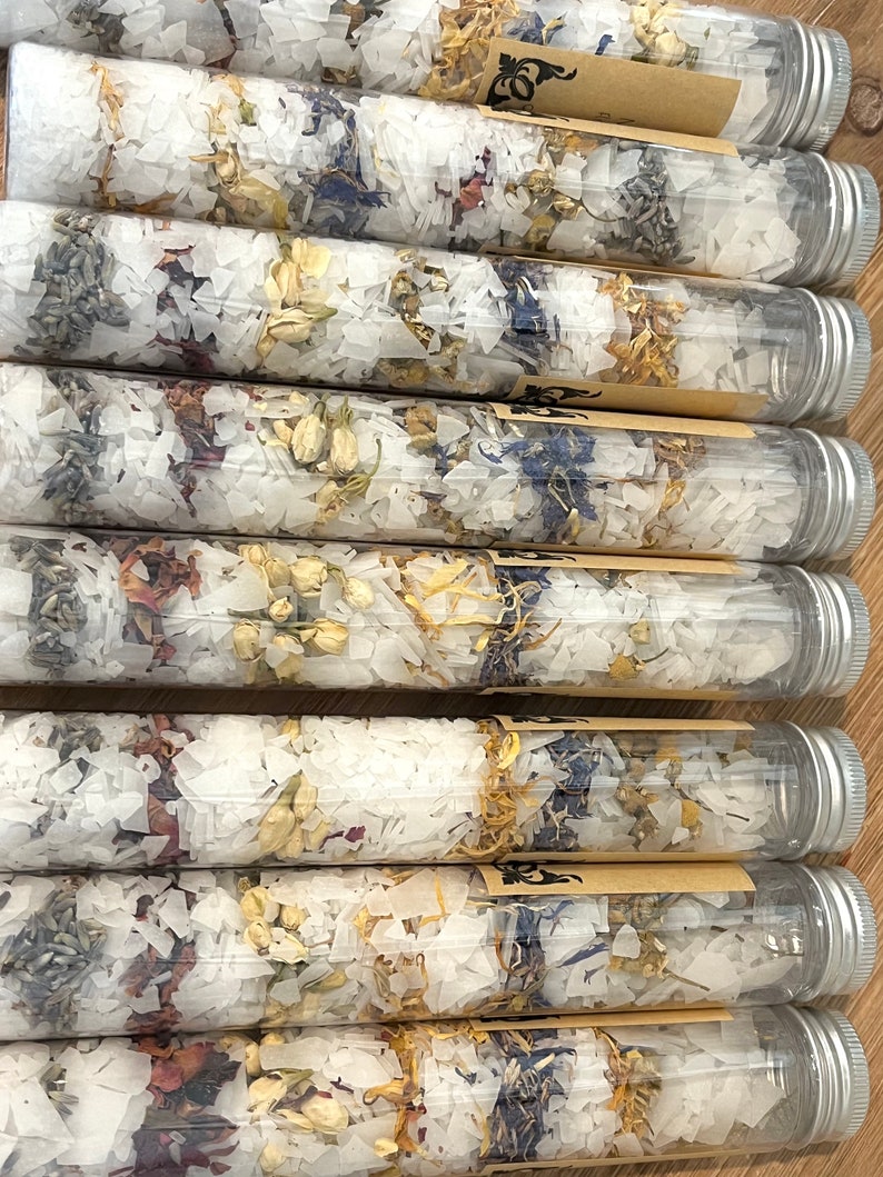 Puede incluir: Primer plano de varios tubos de vidrio transparente llenos de sales de ba&ntilde;o blancas y p&eacute;talos de flores secas. Los tubos tienen tapas plateadas y est&aacute;n dispuestos en fila.