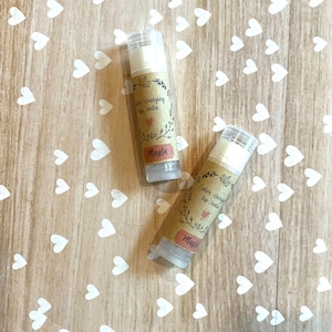 May include: Two colour changing lip balms with brown labels that say "Colour Changing Lip Balm" and "Mayla". The lip balms are on a wooden surface with white hearts.