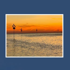 May include: A serene seascape at sunset. The sky is ablaze with orange and yellow hues, reflecting on the calm water. Several dark, slender poles with small, diamond-shaped signs stand in the water, silhouetted against the vibrant sky.