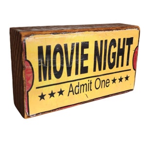 Puede incluir: Un letrero de madera amarillo y rojo con el texto "Movie Night" y "Admit One" con tres estrellas a cada lado del texto.