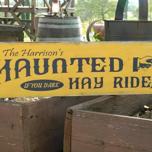 May include: A yellow wooden sign with black text that reads "The Harrison's Haunted Hay Rides If You Dare" with a black silhouette of a hand pointing to the right.