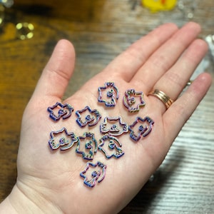 May include: A collection of small, iridescent cat-shaped charms. The charms are held in a hand and display a range of colours, including blue, purple, and green. The charms have a cutout design and are likely used for jewellery or crafting.