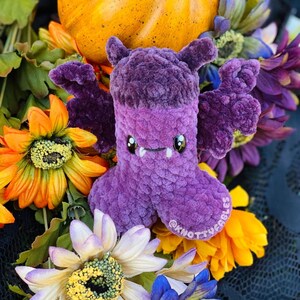 Peut inclure: Chauve-souris en peluche violette avec un sourire narquois et de grandes oreilles tombantes. La chauve-souris est entour&eacute;e de fleurs aux couleurs automnales.