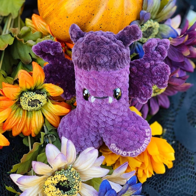 Peut inclure: Chauve-souris en peluche violette avec un sourire narquois et de grandes oreilles tombantes. La chauve-souris est entour&eacute;e de fleurs aux couleurs automnales.