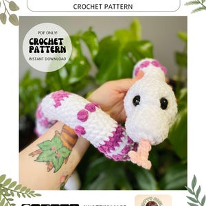 May include: Crochet pattern for a white and purple snake named Vaelis. The pattern is available as an instant download. The image shows a crocheted snake with pink accents and black eyes.