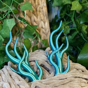 Peut inclure: Deux sculptures de plantes stylis&eacute;es, de couleur turquoise, avec des tiges incurv&eacute;es et des pointes, nich&eacute;es dans un tissu beige doux. L'arri&egrave;re-plan pr&eacute;sente un feuillage vert luxuriant et une texture tiss&eacute;e.