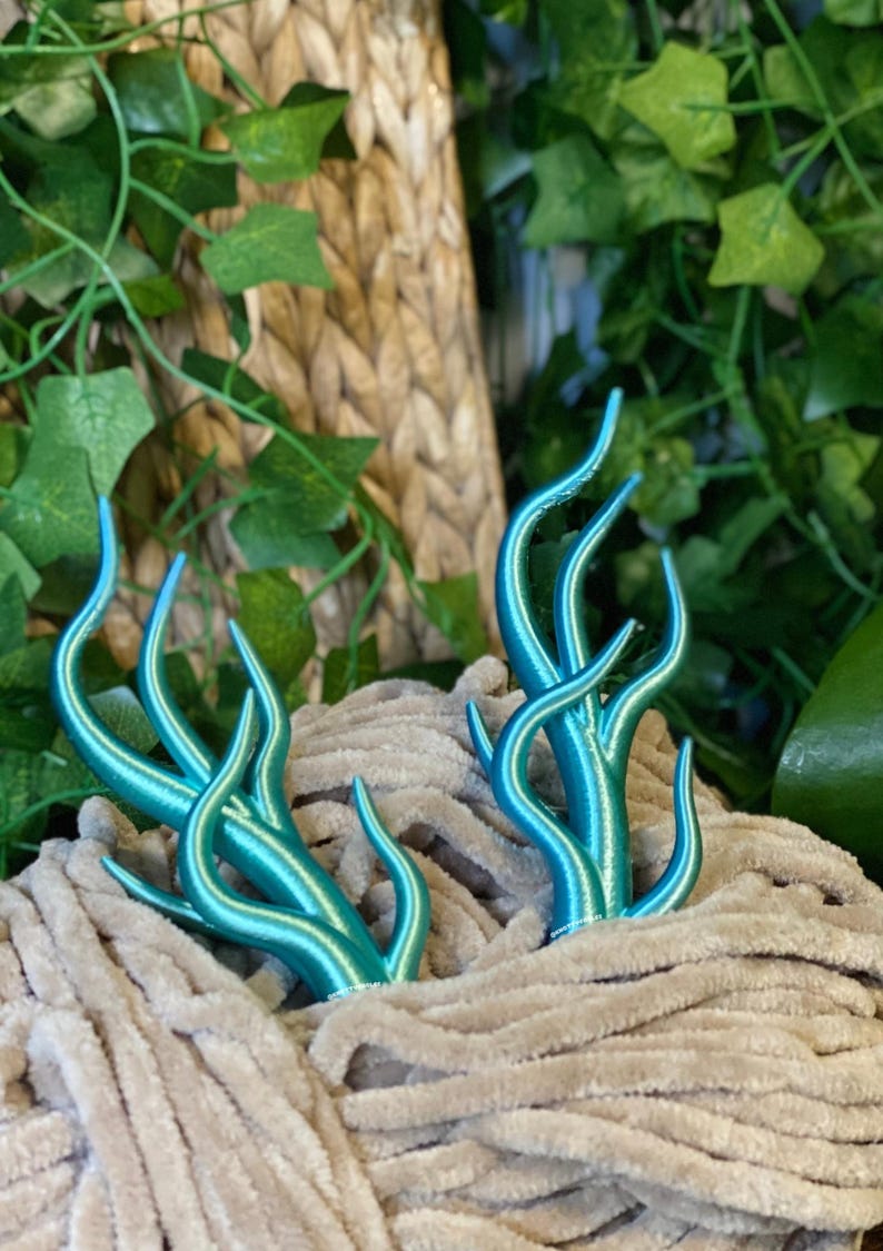 Peut inclure: Deux sculptures de plantes stylis&eacute;es, de couleur turquoise, avec des tiges incurv&eacute;es et des pointes, nich&eacute;es dans un tissu beige doux. L'arri&egrave;re-plan pr&eacute;sente un feuillage vert luxuriant et une texture tiss&eacute;e.