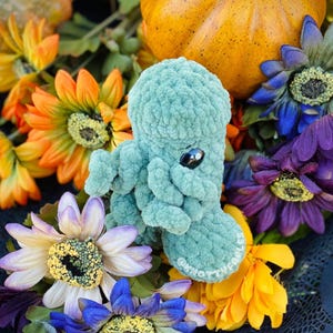 Peut inclure: Un jouet en peluche en forme de pieuvre crochet&eacute; bleu turquoise avec des yeux noirs, entour&eacute; de fleurs artificielles color&eacute;es.