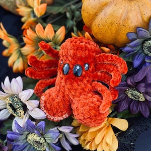 Peut inclure: Une araign&eacute;e orange en crochet avec de grands yeux noirs, entour&eacute;e de fleurs d'automne. L'araign&eacute;e est faite avec un fil pelucheux et a une apparence textur&eacute;e.