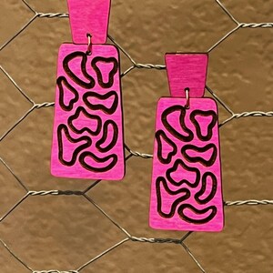 May include: A pair of pink wooden earrings with a geometric pattern. The earrings are rectangular in shape and have a black outline.
