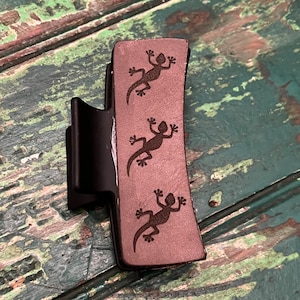 May include: A brown plastic hair clip with three black lizard silhouettes engraved on the front.