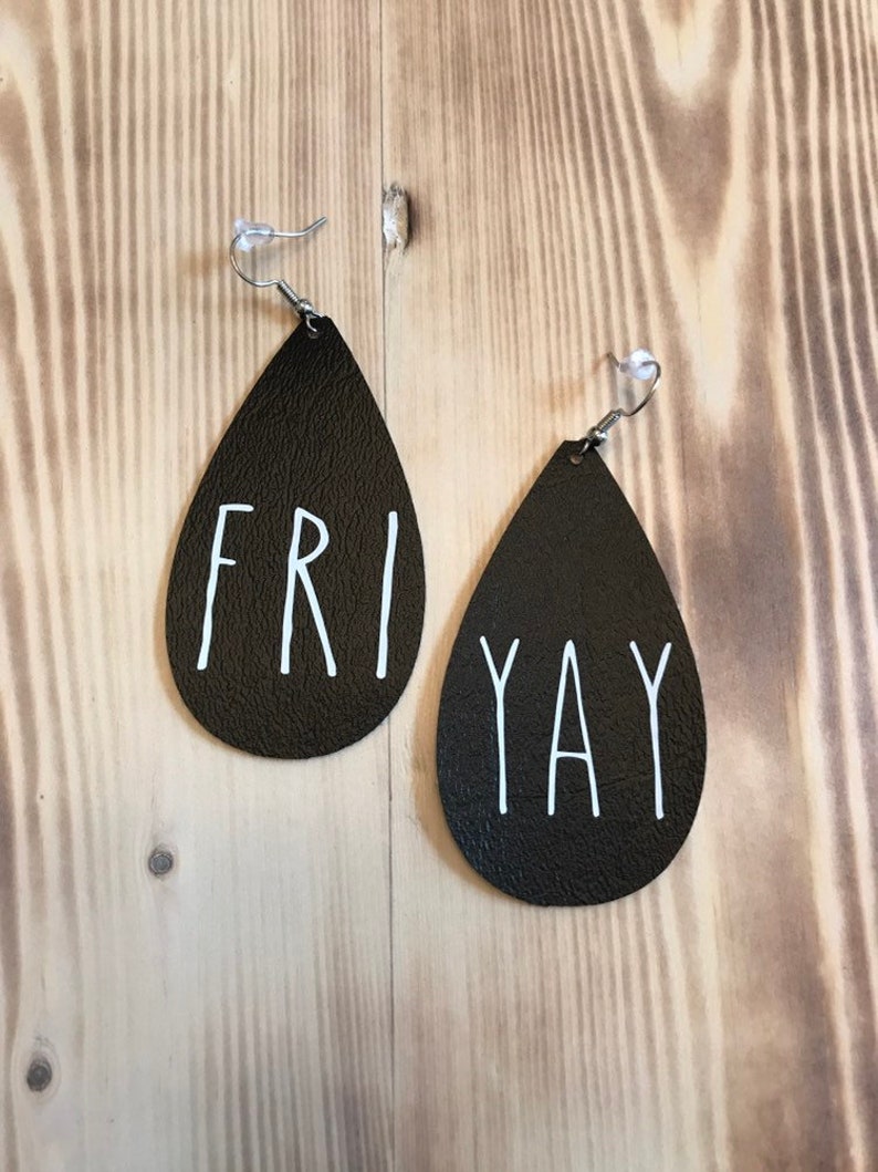 Puede incluir: Un par de pendientes de cuero negro en forma de l&aacute;grima con letras blancas. El pendiente izquierdo dice "FRI" y el derecho dice "YAY".