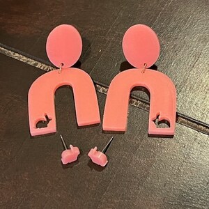 Op de afbeelding: Een paar roze statement oorbellen met een konijnen-silhouet uitsnijding. De oorbellen zijn boogvormig en hebben een kleine roze knop.