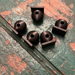 Può includere: Una collezione di piccoli oggetti sferici neri con un foro centrale, ciascuno attaccato a una base rettangolare. Gli oggetti sono disposti su una superficie di legno verniciata di verde e usurata.