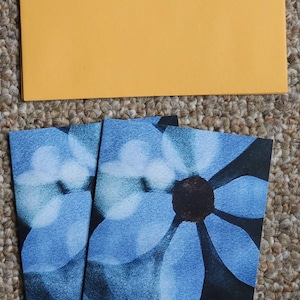 May include: An overhead shot of stationery. A mustard yellow envelope is on top, with two greeting cards below. The cards feature a blue floral design on a black background. The cards are folded.