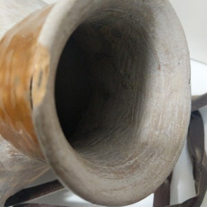 May include: Close-up of a weathered, light-colored drum with a wide opening. The drum has a brown leather strap and a warm, golden-brown band around the body. The interior is dark, contrasting with the lighter exterior.