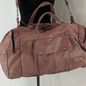 May include: A dusty rose leather duffel bag with a zipped pocket on the front and a top handle. The bag has a shoulder strap and is made of a soft, supple leather.