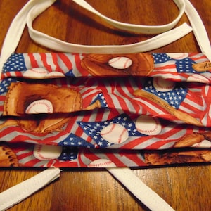 May include: A white fabric face mask with a red, white, and blue American flag pattern. The pattern also includes baseballs, bats, and baseball gloves. The mask has white ties.