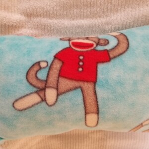 May include: A rectangular, light blue fleece pillow featuring a cartoon sock monkey design. The monkey wears a red shirt and has brown limbs and a white face. The pillow is a soft, plush item.