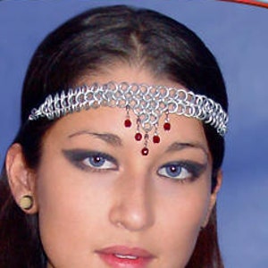 May include: A woman wearing a silver chainmail headband with red beads and a silver chainmail necklace with a silver pendant. She is wearing a black top and has long dark hair.