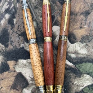 May include: Three wooden pens with different wood grain patterns and metal accents. The pens have a gold-colored tip and a silver-colored band.
