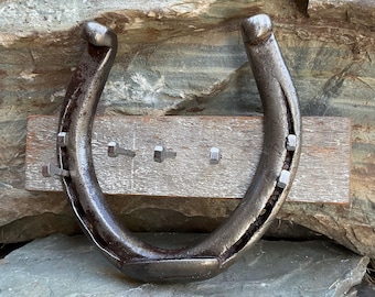 May include: A rustic key holder made from a horseshoe mounted on a wooden board. The horseshoe is made of metal and has a weathered finish. The board is made of wood and has a natural finish. There are five metal hooks on the board for hanging keys.