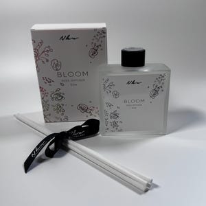 May include: A rectangular frosted glass reed diffuser bottle with a black cap and a matching white box, both labeled Bloom Reed Diffuser 6.7oz with floral line art. A bundle of white reeds tied with a black ribbon featuring white text is included.
