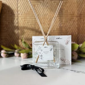 May include: A clear glass diffuser bottle with wooden sticks and a white label that reads "Bloom Scented Diffuser 200 ml". The bottle is sitting on a white surface with a floral design on the label.