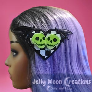 May include: A black and white checkered heart-shaped hair clip with two green skull faces and black bat wings. The clip is on a mannequin with purple hair.
