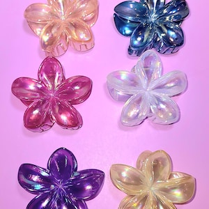 May include: Six iridescent flower-shaped hair clips in various colours, including pink, purple, blue, clear, and yellow. The clips are arranged in two rows of three on a pink background.