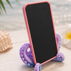 May include: A smartphone with a coral pink case resting in a purple octopus tentacle stand. The stand features detailed suction cups and a curved design that holds the device upright. The phone screen is black and reflects light from the surroundings.