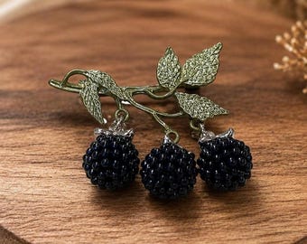 May include: A brooch featuring a textured green metal branch with detailed leaves and three dangling dark blue beaded blackberry charms. The berries are composed of small, round, dark blue beads clustered together to resemble the fruit's surface.