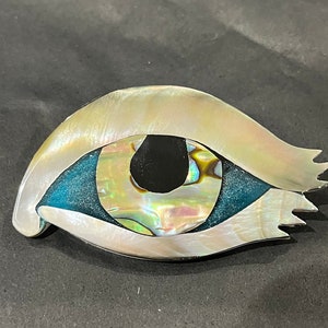 Puede incluir: Un broche blanco, iridiscente, en forma de ojo con un centro azul y verde. El broche está hecho de nácar y tiene un borde dentado.