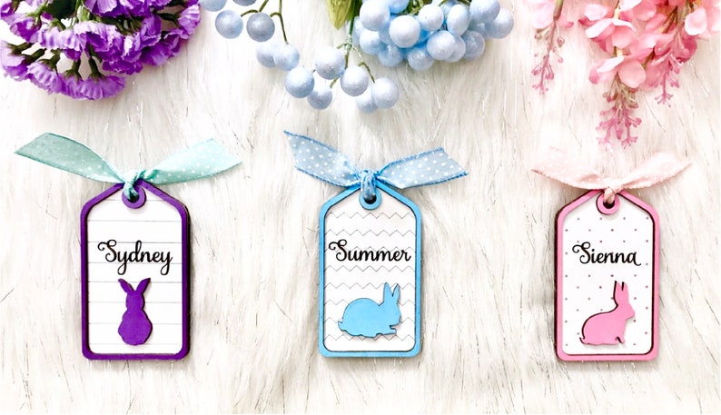 May include: Three wooden gift tags with bunny silhouettes and personalized names: Sydney, Summer, and Sienna. Each tag is decorated with a different pattern and color: purple, blue, and pink.