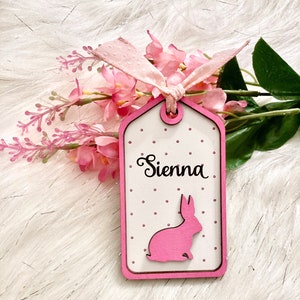 May include: A pink wooden tag with white polka dots and a pink bunny silhouette. The tag is tied with a pink ribbon and says "Sienna" in cursive.