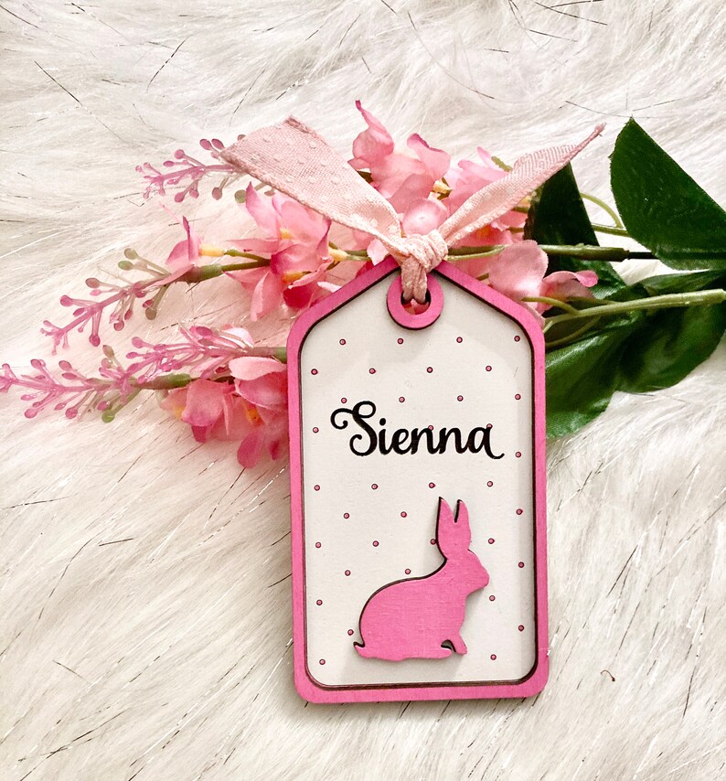 May include: A pink wooden tag with white polka dots and a pink bunny silhouette. The tag is tied with a pink ribbon and says "Sienna" in cursive.